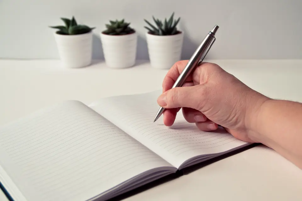 A hand writing with a pen in an open notebook on a white desk